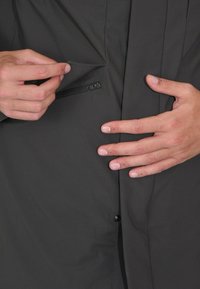 Camicia grigia scura con bottoni e tasca con cerniera nascosta. Il tessuto appare liscio con una texture minima e il design è essenziale.