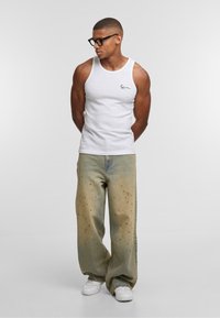 Muškarac u crnim naočalama, bijeloj majici bez rukava, labavim izblijedjelim trapericama sa mrljama boje i bijelim tenisicama, stoji s rukama iza leđa.