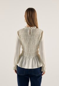 Witte peplum blouse met gathered textuur, hoge hals en sleufdetail aan de achterkant. Lange mouwen met elastische manchetten. Draagt bij donkerblauwe jeans.
