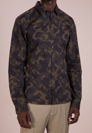 Mann mit einem dunkelgrünen und schwarzen Camouflage-Button-up-Hemd mit langen Ärmeln und beiger Hose, stehend vor einem schlichten Hintergrund.