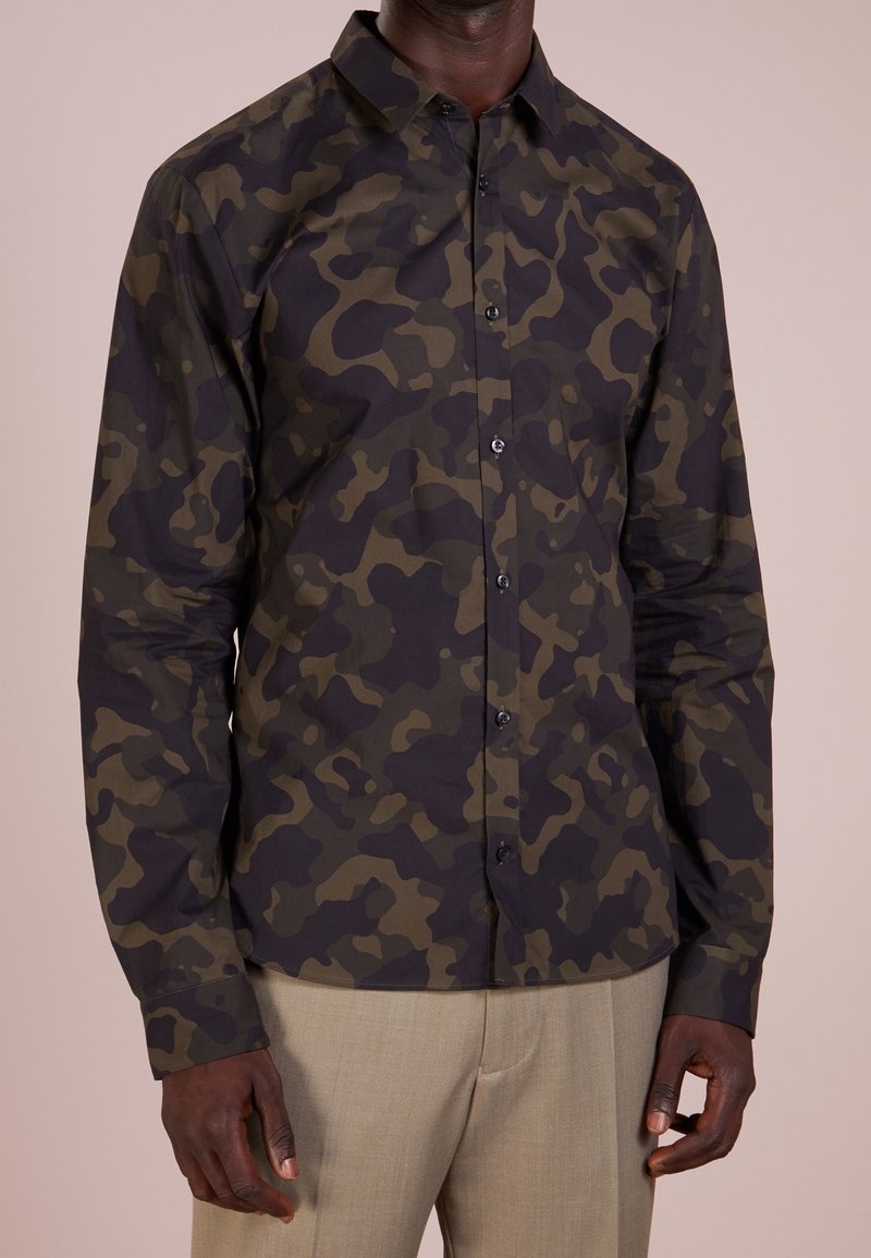Homme portant une chemise à manches longues boutonnée à motif camouflage vert foncé et noir, avec un pantalon beige, debout devant un fond uni.
