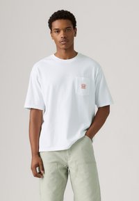 White short-sleeve T-shirt made of cotton, featuring a chest pocket with a red label. Loose fit, round neckline, paired with light green pants.