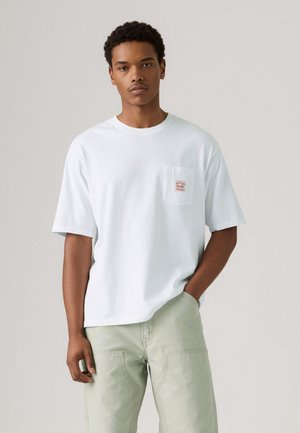 White short-sleeve T-shirt made of cotton, featuring a chest pocket with a red label. Loose fit, round neckline, paired with light green pants.