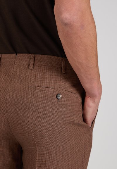 Close-up of a person wearing brown checkered pants with a buttoned back pocket and a black shirt tucked in.