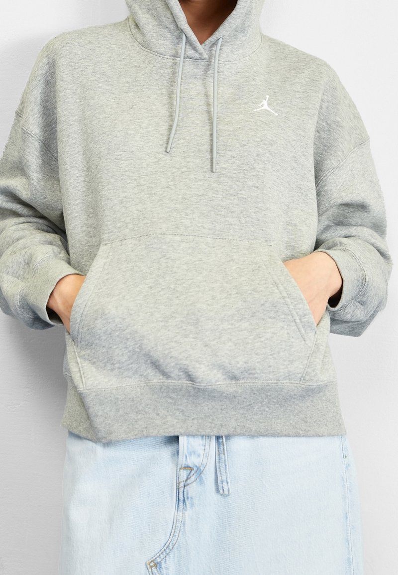 Personne portant un sweat à capuche gris clair avec un logo blanc, les mains dans la poche avant, assorti à un jean en denim bleu clair.