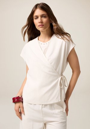 Top avvolgente bianco in tessuto leggero con maniche corte e un nodo in vita, sovrapposto a una camicia con dettagli di borchie decorative.