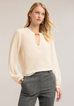 Blond vrouw die een crèmekleurige blouse draagt met een gouden kettingdetail en grijze krijtstreeppantalon, staand met één hand in de zak tegen een neutrale achtergrond.