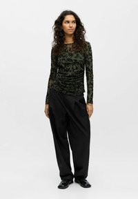 Long-sleeve fitted top in dark olive green with black floral pattern, paired with high-waisted black wide-leg trousers and glossy black shoes.