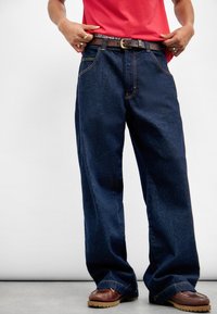 Mørkeblå vide denimjeans med kontrasterende oransje sømmer, metallglidelås og et brunt lærbelte med spenne.