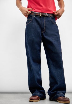 Jean large bleu marine en denim, avec des surpiqûres orange contrastantes, une fermeture éclair en métal et une ceinture en cuir marron avec une boucle.