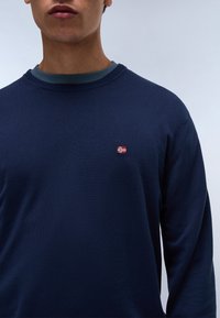 Marineblauwe trui met een ronde hals, voorzien van een groene binnenkraag en een klein rood-wit vlaggenembleem op de borst.