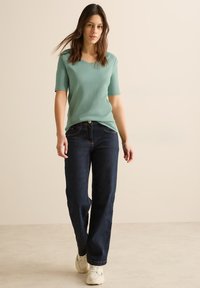 Mintově zelený top s krátkým rukávem a véčkovým výstřihem, spárovaný s tmavě modrými širokými kalhotami a bílými teniskami, stojící na neutrálním pozadí.