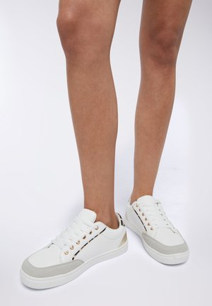 Sneakers bianchi a basso profilo con punta in suede grigio, occhielli dorati e un accento metallico sul tallone, posati su gambe di colore chiaro.