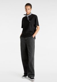 T-shirt en coton noir à manches courtes, avec un petit logo. Associé à un pantalon ample gris foncé et une écharpe à carreaux autour du cou.