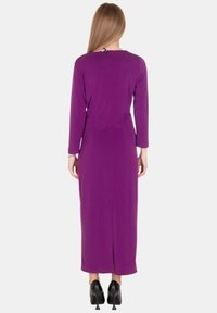 Abito lungo viola con maniche lunghe, silhouette aderente, dettaglio spacco posteriore e scollatura rotonda, abbinato a scarpe nere con tacco alto.