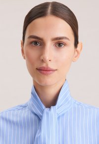 Hellbluse mit Kragen in blauen Farbton und weißen vertikalen Streifen, mit einer gebundenen Detail am Hals. Der Stoff wirkt glatt und strukturiert.