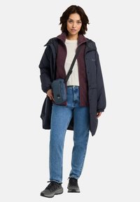 Cappotto lungo blu scuro su una giacca in pile bordeaux, camicia chiara, jeans blu e scarpe grigie; porta una piccola borsa a tracolla blu.