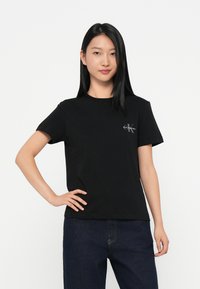 Melna kokvilnas T-krekls ar īsām piedurknēm, apaļu kakla izgriezumu un nelielu baltu logo kreisajā krūšu daļā. Vienkāršs, bezšuvju dizains.