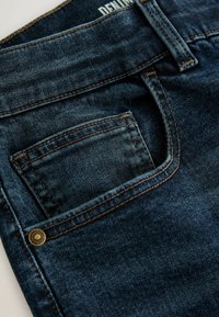 Dark blue denim jeans featuring visible stitching, a back pocket with a brass rivet, and a visible brand label at the waistband.