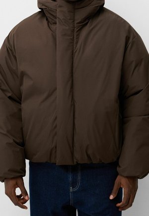 Homme portant une veste matelassée marron foncé avec un col montant et un ourlet élastique, associée à un jean bleu foncé, visible du cou à la taille.