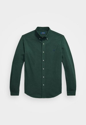 Mörkgrön långärmad skjorta med knappar, knäppt krage och liten broderad logotyp på vänster bröst mot vit bakgrund.