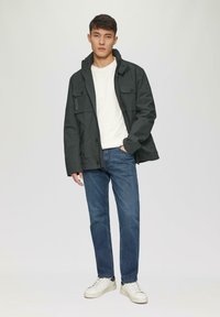 Dunkelgrüne Jacke mit einem hohen Kragen, mehreren Fronttaschen und einem Reißverschluss, kombiniert mit blauen Jeans und weißen Sneakern.