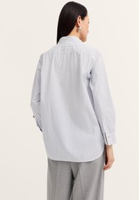 A woman with long dark hair wears a light blue and white striped shirt, viewed from the back, with gray slacks and large silver earrings.
