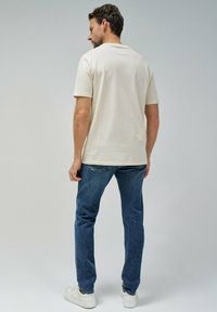 Camiseta de manga corta color crema con escote redondeado, combinada con vaqueros azules y zapatillas blancas, vista desde la parte de atrás.