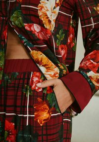 Floral-print garment in burgundy with plaid pattern, featuring vibrant blossoms and contrasting cuffs, with a pocket detail and soft texture.