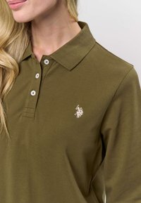 Oliivinvihreä poolopaita, joka on valmistettu pehmeästä kankaasta. Siinä on klassinen kaulus, kolme valkoista nappia ja pieni kirjailtu logo rinnassa.