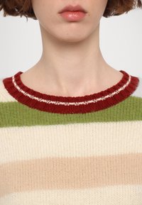 Gros plan d'une personne portant un pull en laine tricoté avec des rayures horizontales vertes, beiges et blanc cassé, et un col côtelé rouge.