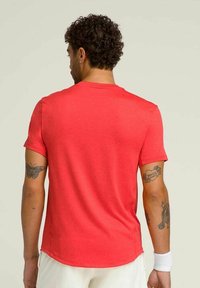Camiseta roja de manga corta con cuello redondo, textura suave y dobladillo curvado. Presenta tatuajes en los brazos visibles.