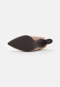 Bota de tacón de ante beige con puntera afilada, con textura negra antideslizante en el tacón y la suela, descansando sobre una superficie blanca.