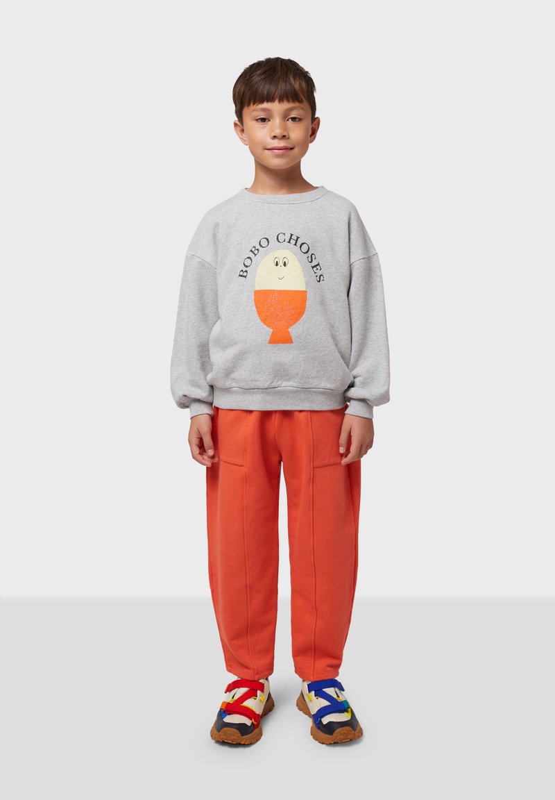 Sudadera gris con un gráfico de caricatura y el texto "BOBO CHOSES". Pantalones naranja brillante, sandalias coloridas con tiras rojas, azules y amarillas, y suelas texturizadas.