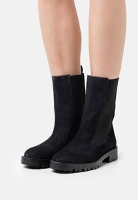 Black suede mid-calf boots with chunky rubber soles worn on bare legs, photographed on a white background.