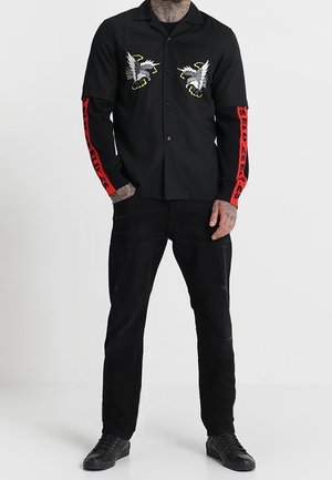 Mand iført sort skjorte med knapper og hvide ørnedesign på brystet samt rødt bogstavtryk på ærmerne, kombineret med sorte bukser og sorte sko.