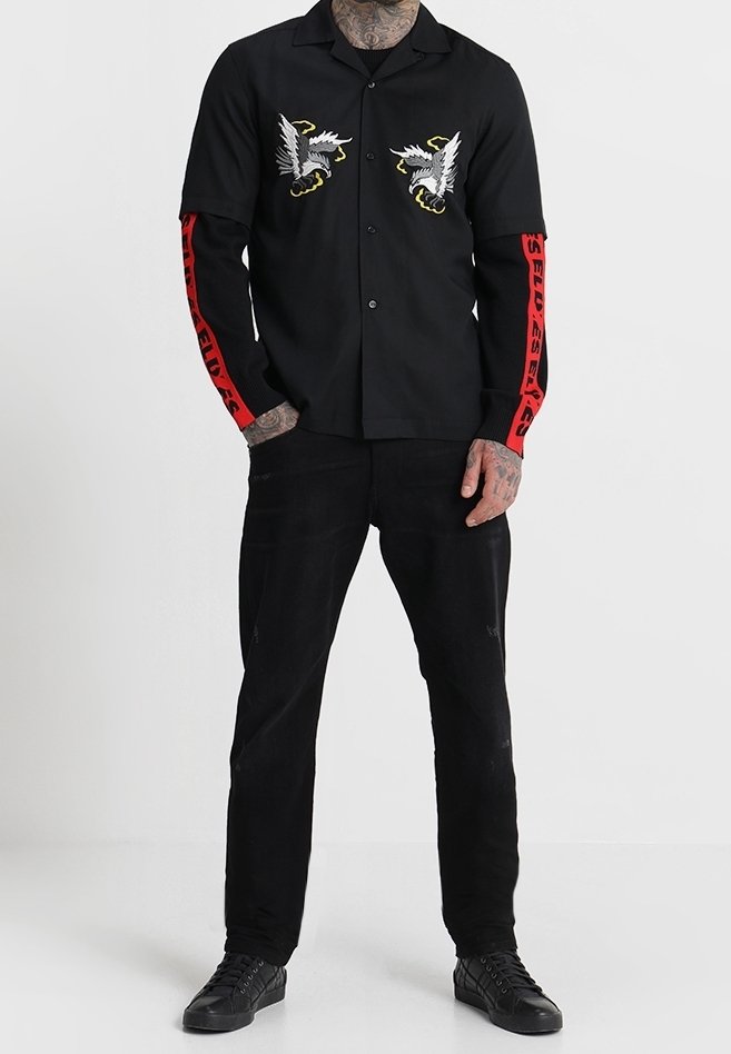 Homme portant une chemise noire boutonnée avec des motifs d'aigle blanc sur la poitrine et des lettres rouges sur les manches, associée à un pantalon noir et des chaussures noires.