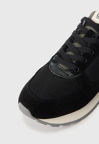 Schwarzer Sportschuh mit einem Obermaterial aus Mesh, Wildlederakzenten, schwarzen Schnürsenkeln und einer grauen Gummisohle. Olivgrüne Details an der Zunge.
