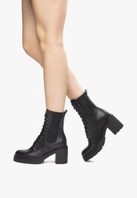Bottines en cuir noir avec un talon épais, devant à lacets et fermeture éclair sur le côté. Présente des coutures texturées et des panneaux élastiques latéraux.