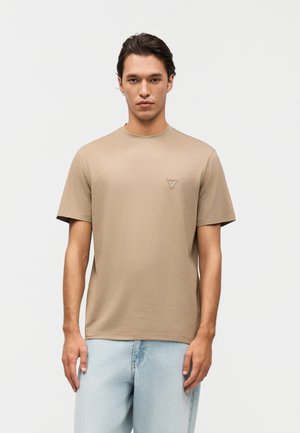 Mann trägt ein beiges T-Shirt mit kurzen Ärmeln und Rundhalsausschnitt, darauf ein kleines dreieckiges Logo auf der Brust, sowie hellblaue Jeans, steht vor weißem Hintergrund.