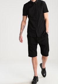 Camicia nera a maniche corte con bottoni e colletto, abbinata a pantaloni corti neri fino al ginocchio e scarpe di pelle nere. Tessuto con una texture liscia.
