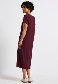 Abito lungo bordeaux a costine con maniche corte, scollatura rotonda e un piccolo dettaglio con bottone sul retro; indossato con scarpe beige senza lacci.