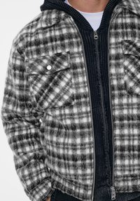 Giacca a quadri grigia e bianca, caratterizzata da un tessuto morbido e strutturato, due tasche sul petto con bottoni e uno strato interno scuro con zip e finiture a coste.