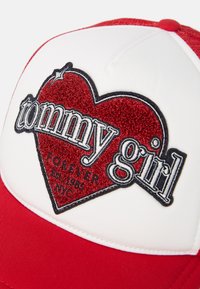 Red and white baseball cap featuring an embroidered red heart patch with “tommy girl” text, black outlines, and decorative accents.