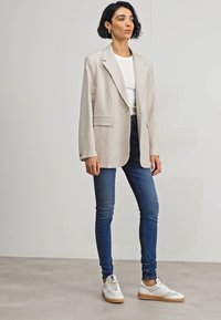 Blazer beige con solapas y bolsillos, camisa blanca, jeans ajustados azules y zapatillas blancas con detalles beiges. Diseño simple y a medida.