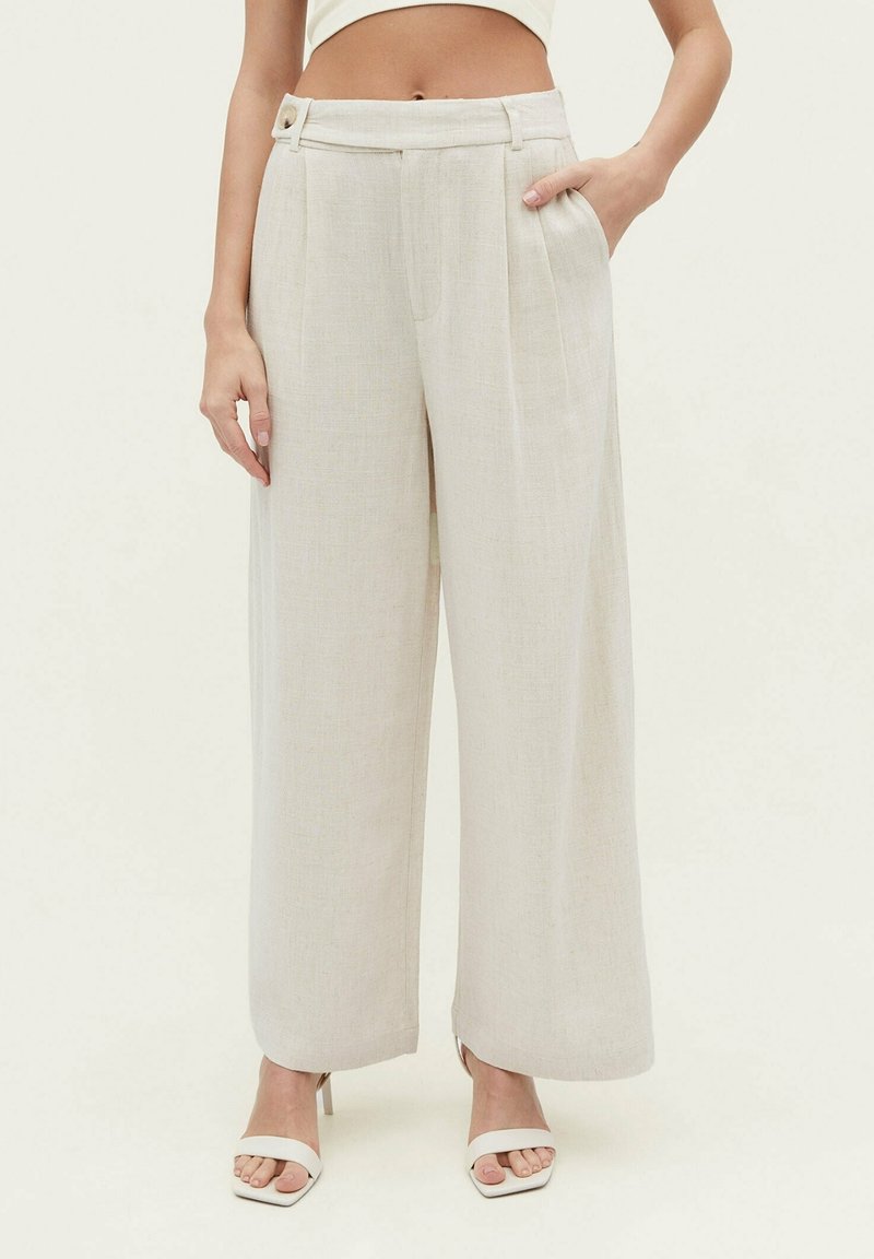 Pantalon à jambes larges en tissu beige clair, avec des plis sur le devant, des poches latérales, un détail bouton à la taille, et une texture lisse.