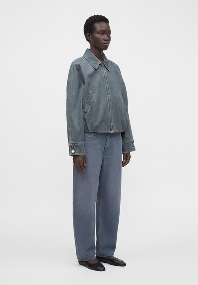 Femme debout portant une veste en denim bleu délavé ample, un jean large assorti et des chaussures plates noires sur un fond uni.