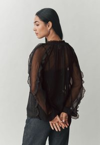 Blusa nera trasparente con maniche a sbuffo e collo alto. Il tessuto presenta righe verticali e una texture leggera e fluida.