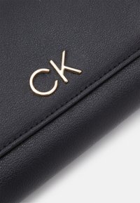 Crna kožna novčanika s teksturiranim površinom, s zlatnim detaljem "CK" logotipa, šivenim rubovima i elegantnim, pravokutnim oblikom.