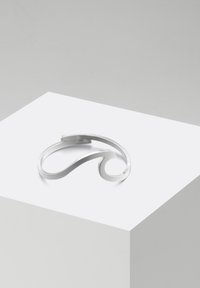 Bracciale a manica in argento con un design curvo e minimalista. Texture metallica liscia, con un accento a forma di onda. Presentato su un piedistallo bianco.
